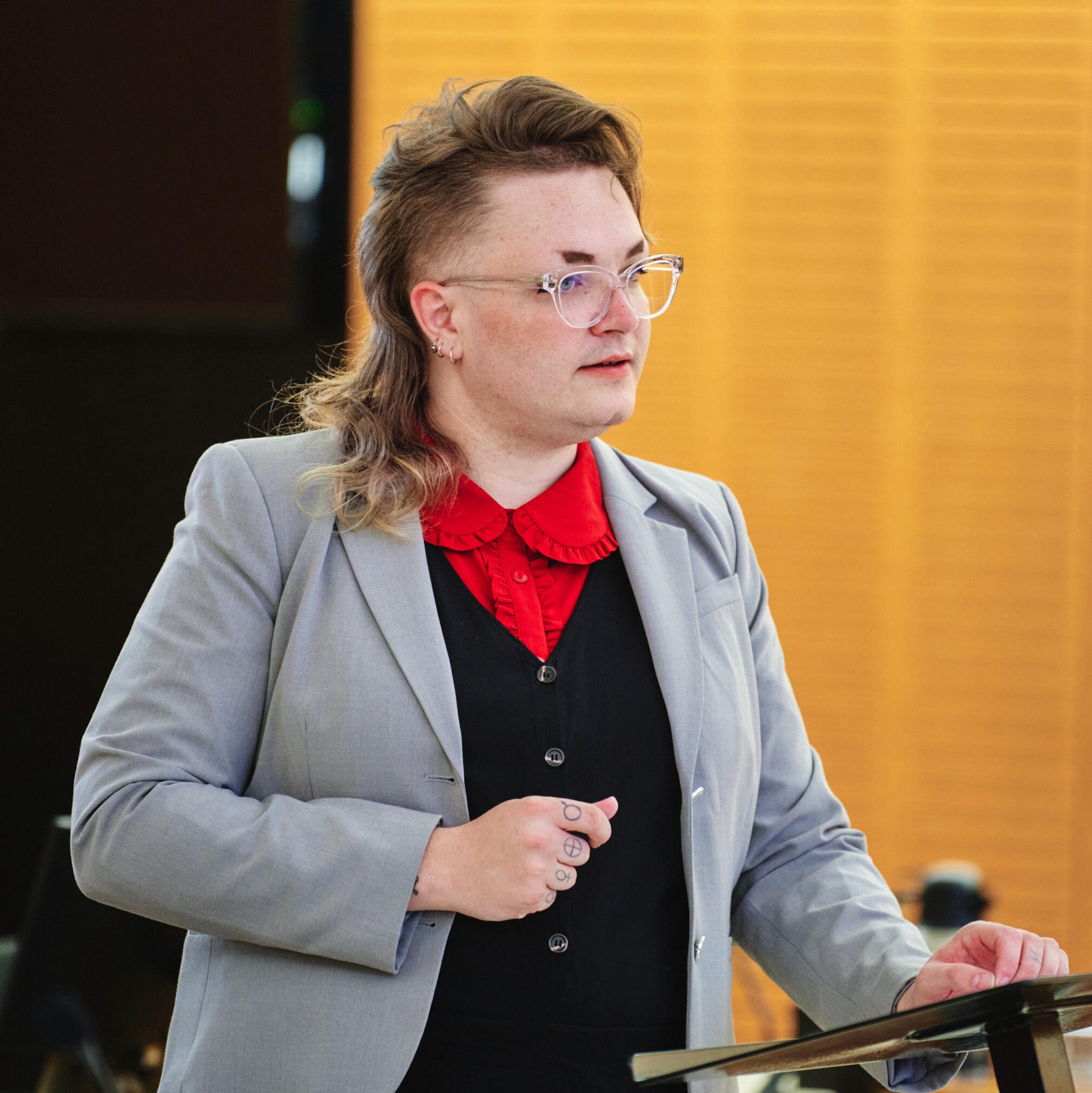 A photograph of Ori Diskett, wearing grey, black and red. She is stood at a bar table lectern.
